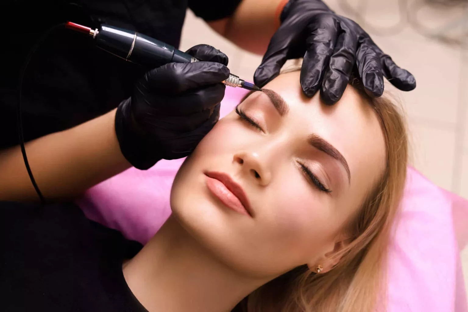 Eine Person mit geschlossenen Augen erhält eine Augenbrauentätowierung. Eine behandschuhte Hand hält eine Tätowiermaschine über der Augenbraue der Person. Die Person liegt auf einem rosa Kissen.